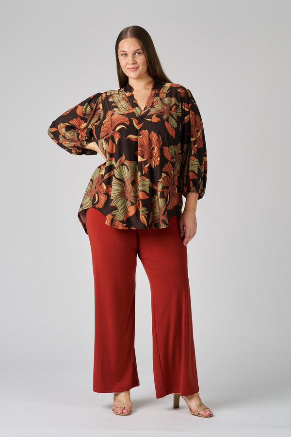 A woman wearing a patterned V-neck top and red pants on a plain background